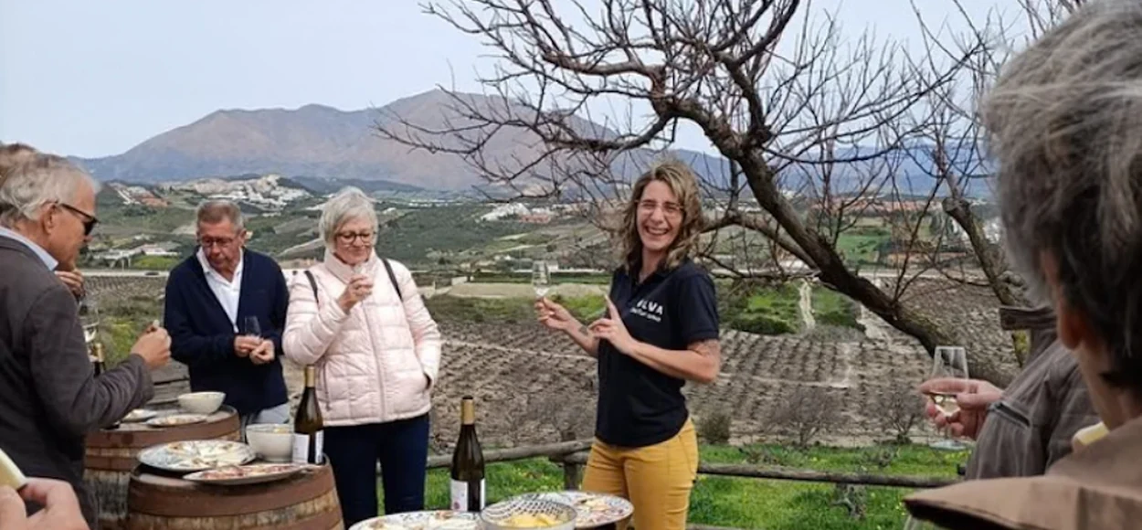 Visitantes disfrutan de una cata de vinos de Nilva entre las viñas de Manilva, junto a barricas utilizadas como mesas.