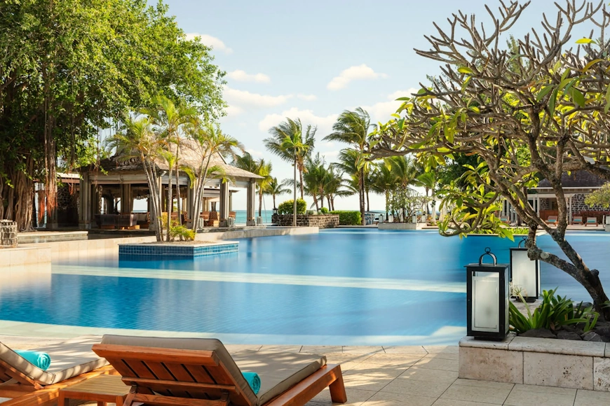Amplia piscina de aguas azules rodeada de palmeras, jardines y tumbonas en The St. Regis Le Morne Resort.