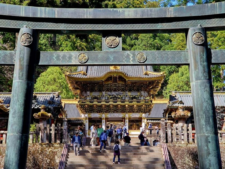 Visitantes subiendo las escaleras del santuario Tōshō-gū de Nikkō, con una puerta monumental de bronce en primer plano y arquitectura ornamentada al fondo.